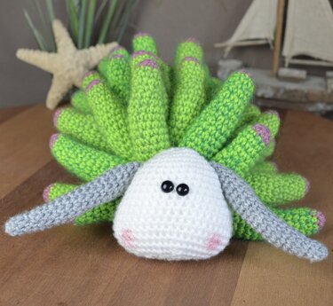 Gehäkelte Amigurumi-Seeanemone mit grünem Körper und weißem Kopf auf Holz.