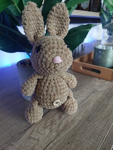 Kuscheliger beiger Amigurumi-Osterhase mit langen Ohren und Knopf am Bauch, vor Pflanzenarrangement