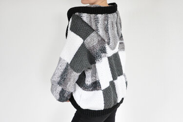 Back view of a person wearing an oversized hooded crochet jacket with a grayscale patchwork of white, gray, and black blocks.