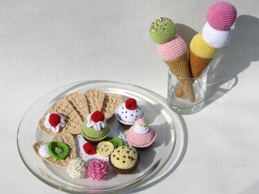 Verschiedene gehäkelte Spiel-Desserts: Eistüten in einem Glas, Cupcakes, Waffeln und Kekse auf einer Glasplatte