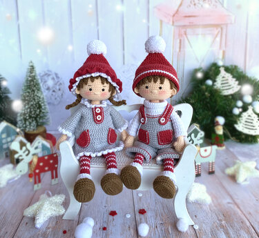 Pair of amigurumi elf dolls in red and grey outfits sitting on a white bench with Christmas decorations.