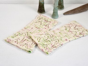 Pair of cream fingerless knitted mitts with green and pink speckles and a lace openwork pattern, laid flat on a white surface.