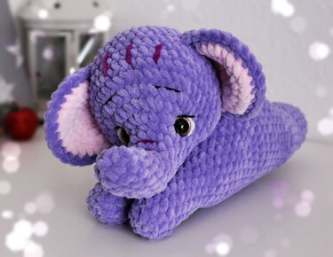 Lila gehäkelter Amigurumi-Elefant mit rosa Ohreninnenflächen, auf heller Fläche liegend.