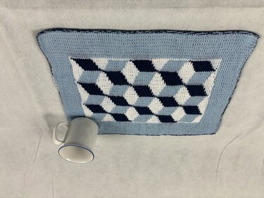 Gestricktes Deckchen in Hellblau, Dunkelblau und Weiß mit 3D-Würfelmuster neben einer Tasse