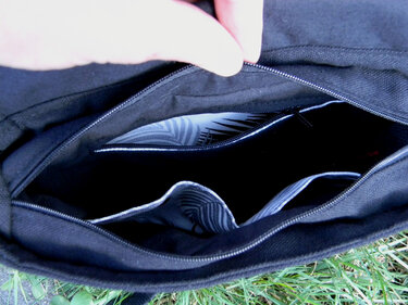 Eine Hand öffnet den Innenraum einer schwarzen Stofftasche, die Reißverschlussfächer und ein gemustertes Futter zeigt.