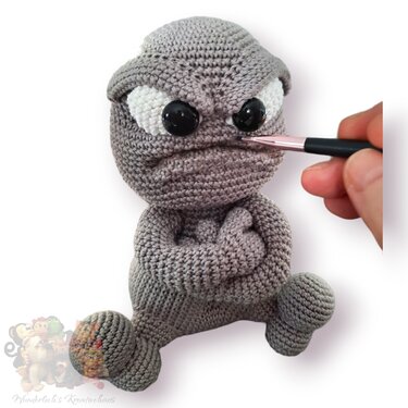 Gray crocheted amigurumi doll with crossed arms and large black eyes being adjusted with a small brush near its nose