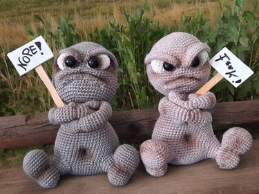 Pair of crocheted grumpy amigurumi figures, one grey and one brown, sitting on a wooden bench holding small signs.