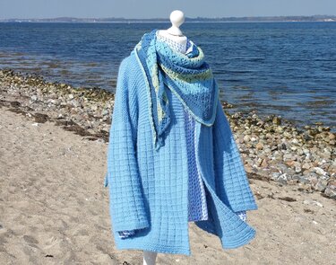 Hellblaue Strickjacke mit Gittermuster und gemustertem Dreieckstuch auf einer Schneiderpuppe am Strand.