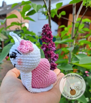 Gehäkelte weiße Amigurumi‑Schnecke mit rosa Herzschale auf Hand vor Gartenpflanzen