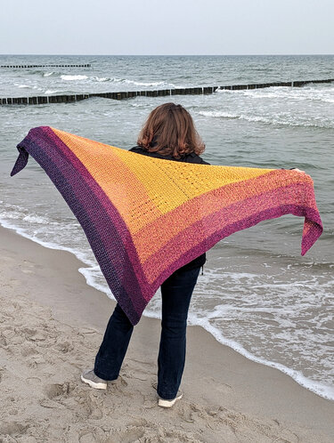 Person am Strand hält gehäkeltes Dreieckstuch in Gelb, Orange, Pink und Lila aufgespannt