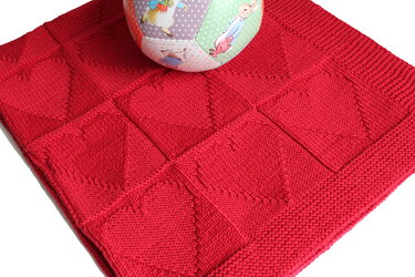 Rote gestrickte Babydecke mit Reliefmuster und buntem Stoffball auf wei
m Hintergrund