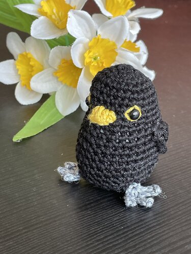 Gehäkelter schwarzer Amigurumi‑Vogel mit gelbem Schnabel vor weißen Osterglocken.