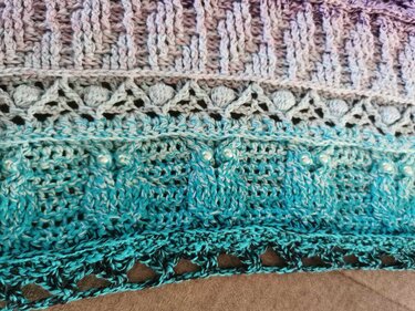 Close-up of a crochet shawl with purple-to-teal gradient, ribbed and lace stitches, and small pearl-like beads.