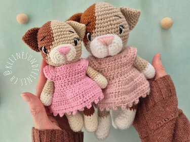 Zwei handgehäkelte Amigurumi-Katzen mit braun-beigem Fell, weißen Pfoten und rosa Kleidern, in Händen gehalten.
