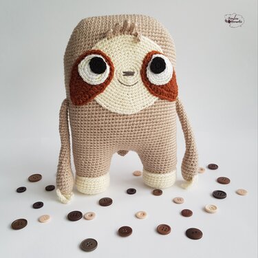 Gehäkeltes beige-braunes Amigurumi-Faultier mit großen Augen vor hellem Hintergrund, umgeben von verstreuten Knöpfen