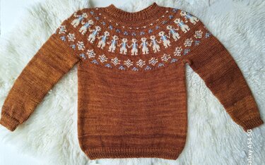 Gestrickter rostbrauner Kinderpullover mit mehrfarbiger Rundpasse aus weißen und hellblauen Figuren- und Schneeflockenmotiven, liegend auf hellem Plüschuntergrund.