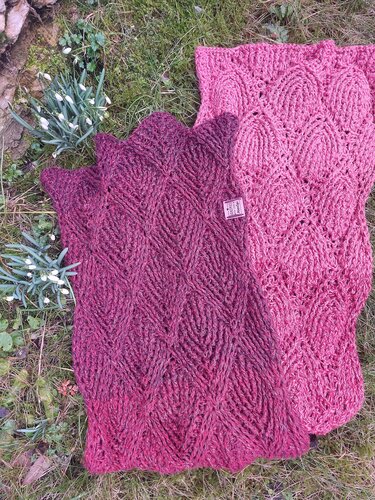 Two crochet scarves, magenta and soft pink, with textured openwork patterns lying on grass near small white flowers