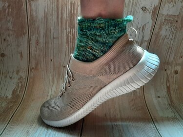 Handgestrickte grüne Socke mit Muster in beigem Sneaker auf Holzuntergrund.