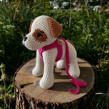 Gehäkelter Amigurumi-Hund weiß und braun mit rosa Geschirr auf Baumstumpf