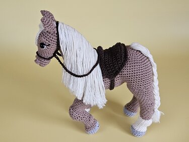 Gehäkeltes Amigurumi-Pferd in Taupe mit weißer Mähne, Schweif, Zaumzeug und braunem Sattel.