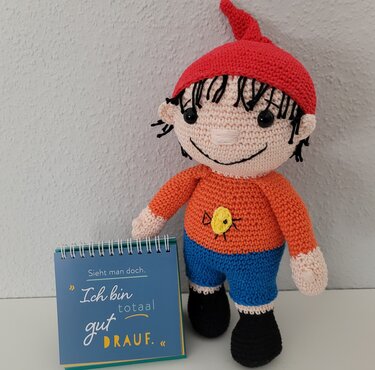 Gehäkelte Puppe mit Zipfelmütze, buntem Outfit und kleinem Buch als Accessoire.