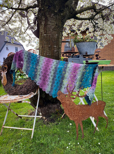 Multicolor gradient crochet triangle shawl with eyelet lace and scalloped edge draped across chairs and a tree in a garden