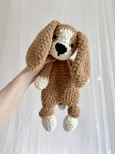 Hand holding a brown and white chunky crocheted puppy amigurumi with long floppy ears against a sheer curtain background with small star accents.