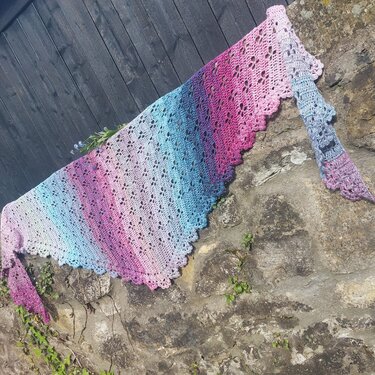 Gehäkeltes Dreieckstuch mit Farbverlauf in Hellblau, Rosa und Violett, drapiert über einer Steinmauer vor einem Holzzaun.