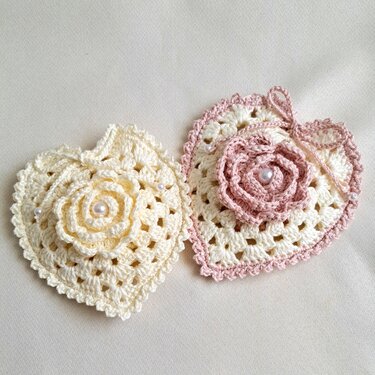 Zwei handgehäkelte Herzapplikationen in Creme und Creme mit rosa Rand, jeweils mit aufgesetzter Häkelrose und kleinen Perlen auf hellem Stoff.