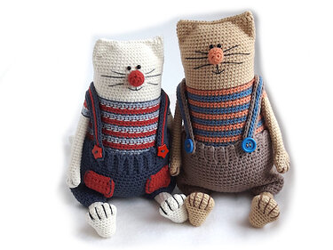 Two crocheted amigurumi cats sitting side by side wearing striped shirts and suspenders with button details