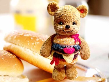 Gehäkelter brauner Teddy mit buntem Burger-Kostüm vor Brötchen