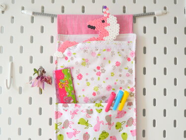 Rosa-weißes Stoff-Utensilo mit Blumen- und Hasenmotiven, hängend an einer weißen Lochwand.
