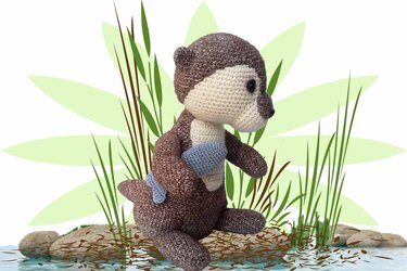 Crocheted brown and cream amigurumi otter holding a small blue fish, seated among reeds