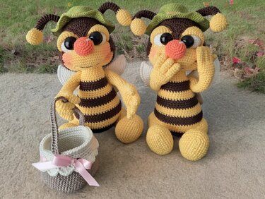 Zwei gehäkelte Bienen-Amigurumi mit gelb-braun gestreiftem Körper, großen schwarzen Augen, grünen Mützchen und kleinem gehäkelten Korb