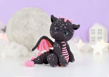 Dark gray amigurumi dragon with pink wings and sparkly speckles sitting in a moonlit setting.