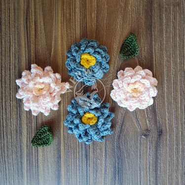 Vier gehäkelte Blumen (zwei blau, zwei rosa) mit gelben Zentren und zwei grüne Blätter auf Holzhintergrund.