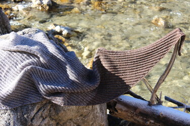 Gestricktes Tuch in Grau-Braun mit Rippenmuster auf Felsen am Bach, mit leicht verschneitem Ast im Vordergrund.
