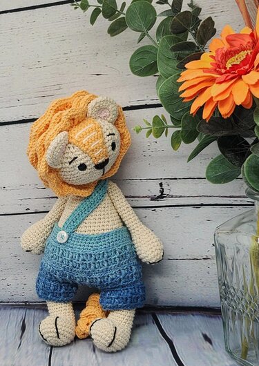 Gehäkeltes Amigurumi-Löwenkind mit gelber Mähne und blauer Latzhose vor hellem Holz-Hintergrund