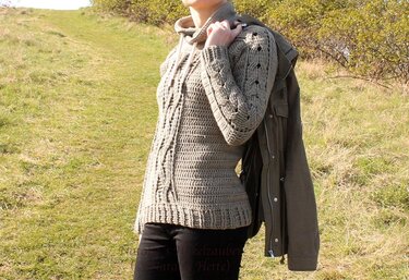 Woman wearing a greige crocheted hooded sweater with cable panels and openwork sleeves, standing on a grassy hill.