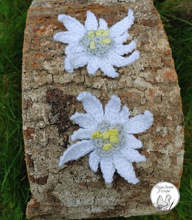 Zwei gehäkelte Edelweißblüten mit weißen Blütenblättern und hellgelb-grauen Zentren auf einem Stück Baumrinde vor Gras.