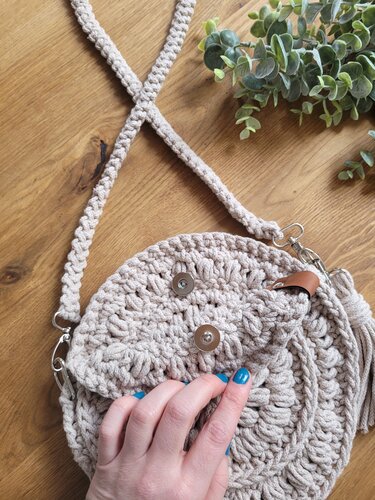 Beige round crocheted shoulder bag with flap and two magnetic snaps, braided strap, and tassel on a wooden surface with a plant.