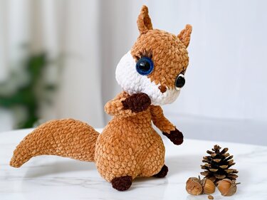 Handmade brown crocheted squirrel amigurumi with white muzzle, large blue and black eyes, standing next to a pine cone and nuts