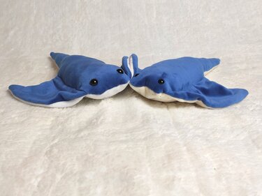 Two blue manta ray plush toys touching noses on a white blanket.
