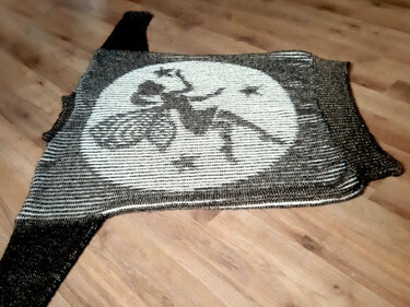 Damen-Strickjacke mit großem Intarsienkreis und Silhouette einer fliegenden Figur auf einem Besen, dunkle und helle Töne, flach auf Holzboden gelegt.