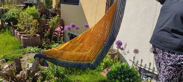 Gehäkeltes Dreieckstuch in Gelb‑Orange zu Blau verlaufend mit Ajourmuster und dunkler Randborte, gehalten im Garten