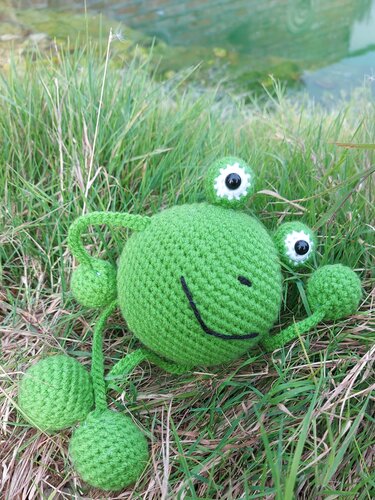 Gehäkelter grüner Amigurumi-Frosch mit großen Augen im Gras am Wasser