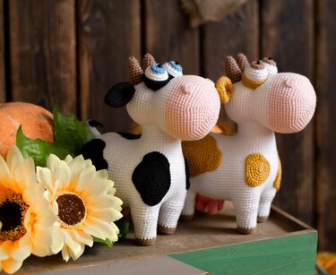 Two crochet amigurumi cow dolls, one black-and-white and one yellow-and-white, beside sunflowers and a pumpkin on a wooden surface.