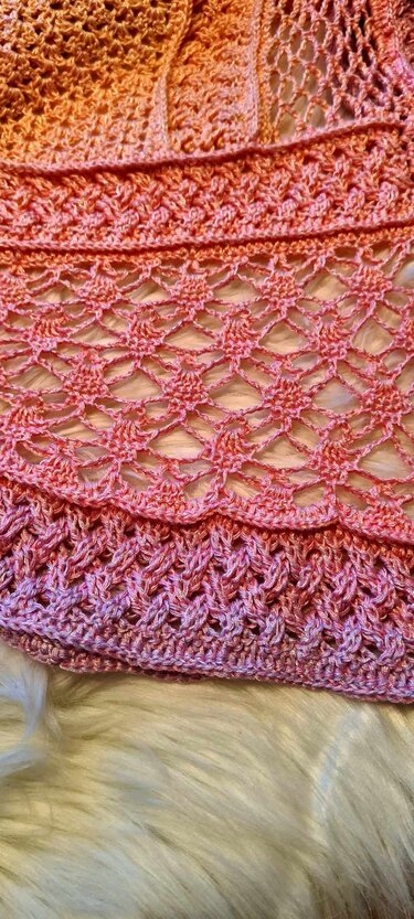 Close-up of a crocheted shawl in peach and pink gradient yarn, showing lace and textured stitch patterns on a soft white surface.