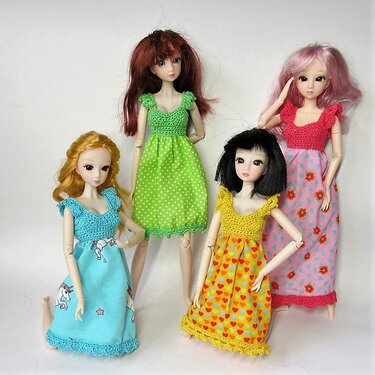 Vier Gelenkpuppen mit unterschiedlichen Frisuren in bunten Kleidern mit gehäkelten Oberteilen und gemusterten Stoffröcken vor hellem Hintergrund.