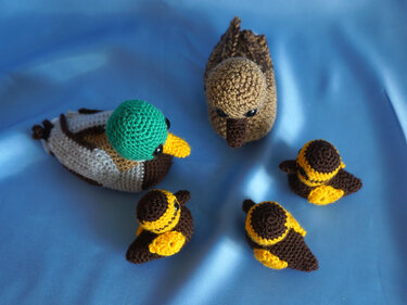 Gehäkelte Amigurumi-Enten: Erpel mit grünem Kopf, braune Ente und drei gelb-braune Küken auf blauem Hintergrund.